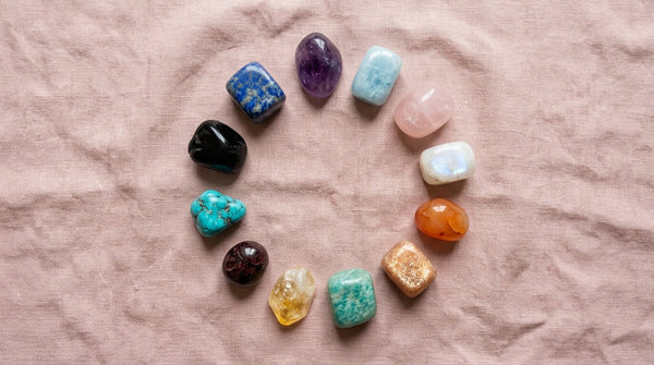 A flat-lay of twelve healing crystals arranged in a circle representing every zodiac sign on a soft pink linen background